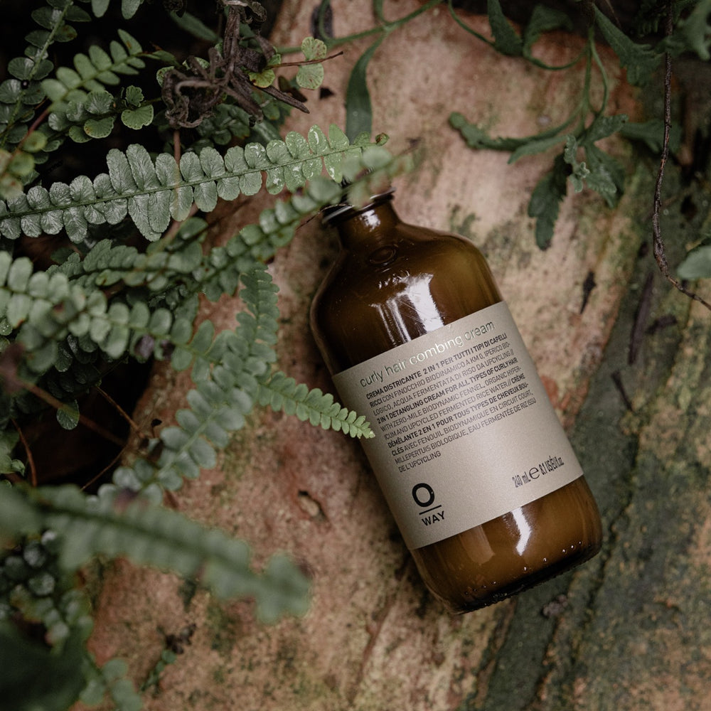 Flacone in vetro da 240ml di curly hair combing cream, crema districante 2 in 1 per tutti i tipi di riccio. Il flacone è poggiato su un tronco in messo al verde delle piante